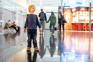 Fototapeta premium Cute baby boy waiting boarding to flight in airport transit hall near departure gate. Active family lifestyle travel by air with children
