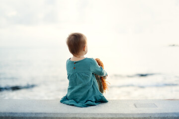 cute european baby toddler in a green dress with a doll sits on a stone bench by the sea, gray background. A girl plays with a doll, a view from the back, sad negative emotions