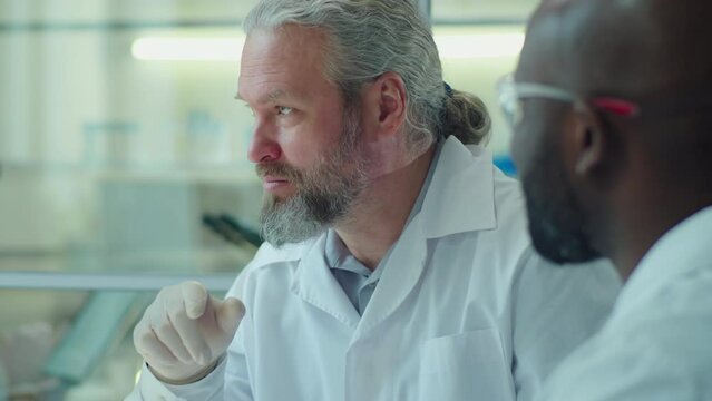 Senior Caucasian scientist in medical gloves and lab coat discussing microscopic image on computer screen with African American colleague