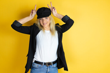 Beautiful woman wearing casual white t-shirt and a cap over isolated yellow background Posing funny and crazy with fingers on head as bunny ears, smiling cheerful