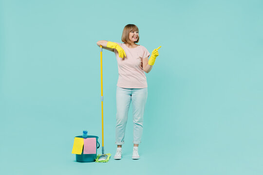 Full body elderly housewife woman 50s in t-shirt gloves doing housework hold mop and bucket point finger aside isolated on plain pastel light blue background. Housekeeping cleaning tidying up concept.
