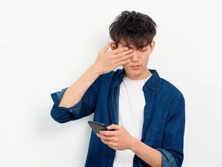 Foto op Plexiglas Portrait of handsome Chinese young man with black curly hair in blue shirt posing against white wall background. Fingers rubbing eyes with mobile phone in hand, looks tired, front view studio shot. © atiger