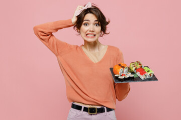 Young mistaken disappointed woman 20s in casual clothes hold in hand makizushi sushi roll served on black plate traditional japanese food scratch hold head isolated on plain pastel pink background
