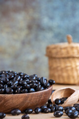 Close-up of wooden bowl with black beans and spoon on wooden table with basket, selective focus, gray background, vertical