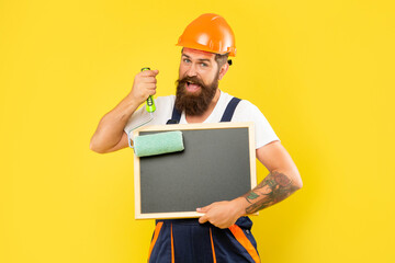 smiling man housepainter in helmet and work clothes hold paint roller and blackboard