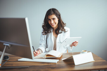 Doctor working at office desk. Female doctor sitting at the table writing in documents and working on laptop computer at office. Female doctor in a modern office clinic / hospital