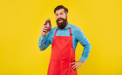 Happy man in red apron holding eggplant yellow background, grocer