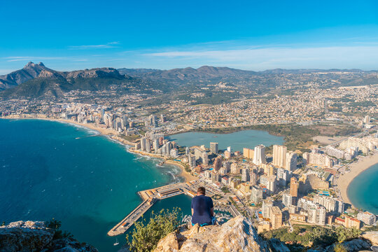 A young man sitting on top of the Penon de Ifach Natural Park in Calpe, Valencia, Valencian Community. Spain. Mediterranean sea. View of the Cantal Roig and La Fossa beach