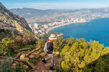 Fototapeta na wymiar A young hiker wearing a hat on the descent path of the Penon de Ifach Natural Park with the city of Calpe in the background, Valencia. Spain. Mediterranean sea. View of La Fossa beach