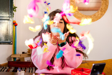 Female handworker blowing bright feathers while sitting at table with supply for making accessories and smartphone and looking at camera