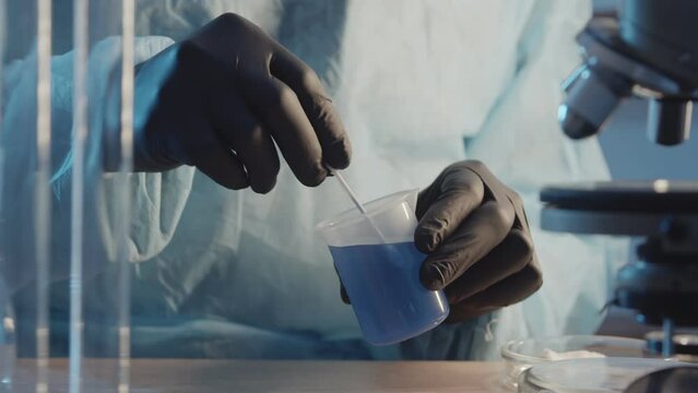 A laboratory assistant in protective rubber gloves stirs a blue liquid in a flask close-up against the background of laboratory equipment. pharmaceutical industry. Research Work