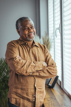 Side view of a happy African retired elderly man standing at home by the window. A satisfied old man looks out the window and smiles