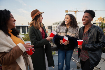 Diverse group of friends standing on rooftop terrace in city, laughing and socializing with friends...
