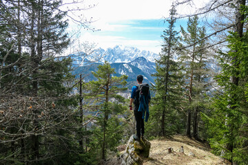 Man with backpack standing on a rock with scenic view of snow capped mountain peaks of Karawanks near Sinacher Gupf in Carinthia, Austria. Mount Wertatscha is visible through dense forest in spring