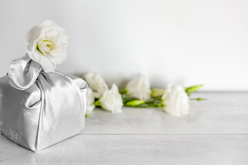 Obraz na płótnie Canvas Gift box wrapped in silk fabric in Furoshiki technique, white flowers Eustoma or Lisianthus, light wooden background.