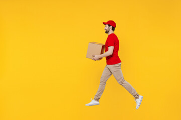 Full body side profile view delivery guy employee man in red cap T-shirt uniform workwear work as dealer courier jump high hold cardboard box run fast jump high isolated on plain yellow background