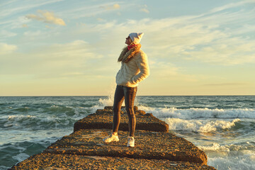A woman with blond hair in a hat and a fur coat walks along the sea along a stone breakwater