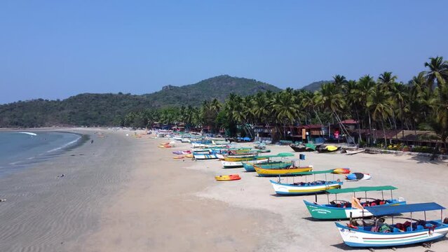 Tourists strolling along the golden sands near the coastline with moored fishing boats in Palolem Beach, in Goa, India - Aerial Fly-over
