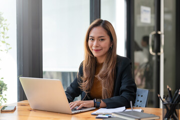 Portrait of smiling young beautiful Asian businesswoman sitting with laptop computer looking at...