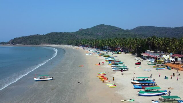 Tourists strolling along the golden sands near the coastline with moored fishing boats in Palolem Beach, in Goa, India - Aerial Fly-over
