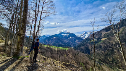 Obraz premium Man with backpack and scenic view of snow capped mountain peaks of Karawanks near Sinacher Gupf in Carinthia, Austria. Mount Hochstuhl (Stol) visible through forest in spring. Rosental sunny day. Awe