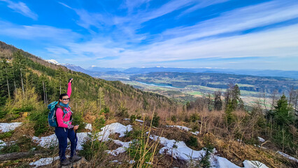Obraz premium Woman with backpack and a scenic view on the Drava river in the Rosental valley on the way to Sinacher Gupf in Carinthia, Austria. Forest in early spring. Austrian Alps mountain ranges on sunny day