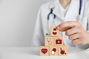 Doctor building pyramid of wooden cubes with different icons at white table, closeup. Insurance concept