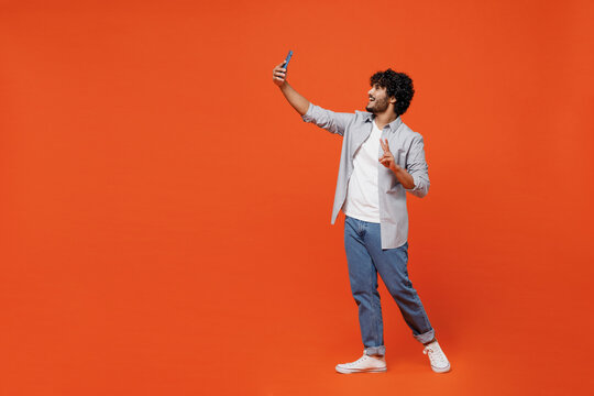Full size body length young bearded Indian man 20s years old wears blue shirt doing selfie shot on mobile cell phone post photo on social network isolated on plain orange background studio portrait.