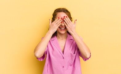 young pretty woman smiling and feeling happy, covering eyes with both hands and waiting for...