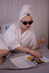 Girl drinking champagne and reading magazine. Attractive woman in bedroom in white bathrobe lies in bed drinking wine and turns over pages of fashion magazine. Close-up view