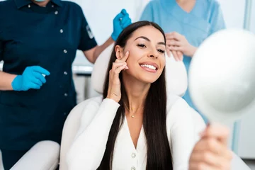 Keuken foto achterwand Schoonheidssalon Beautiful and happy young woman sitting in medical chair and looking in the mirror. She is satisfied after successful beauty treatment with hyaluronic acid fillers or botulinum toxin injections.
