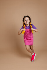 Moving, joyful, little girl touching kanekalon pigtails playing, dancing on one place looking at camera, indulging smiling with missing tooth in pink jumpsuit and purple t-shirt on beige background.