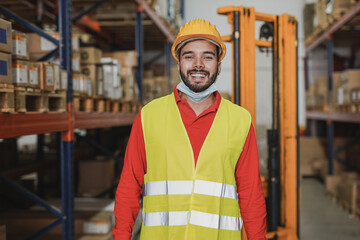 Young warehouse worker man smiling on camera while wearing safety mask under chin