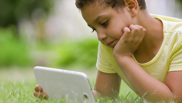 The boy is lying on the lawn with a tablet in his hands. The child uses the gadget in nature. mobile devices are everywhere