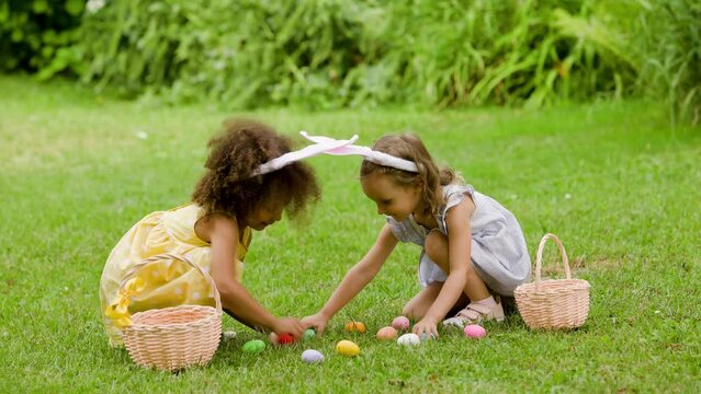 two girls are sitting on the green lawn during easter egg hunt and putting Easter eggs in to baskets