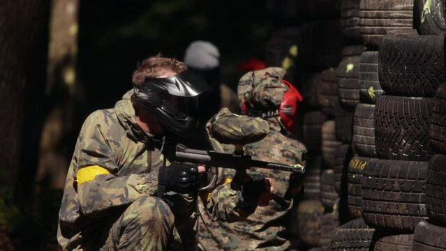 Paintball sport player wearing protective mask aiming gun shelter under gunfire attack with paint splash. Heavily armed masked soldier. Paintball players hide behind tires while shooting paint balls