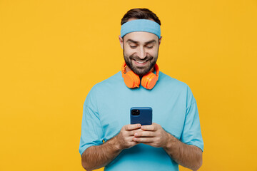 Young fitness trainer instructor sporty man sportsman in headband blue t-shirt spend weekend in home gym hold in hand use mobile cell phone isolated on plain yellow background. Workout sport concept.