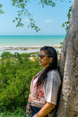 Fototapeta premium Mulher passeando em mirante encostada em árvore, com linda paisagem ao fundo onde vemos o mar, pequeno rio se encontrando com o mar, e céu azul em Cabrália Bahia