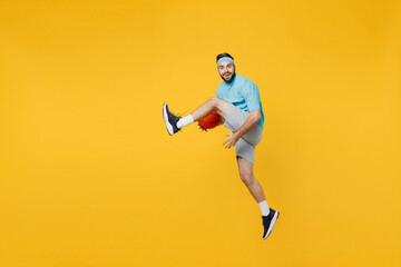 Naklejka na ściany i meble Full body caucasian young fitness trainer instructor sporty man sportsman in headband blue t-shirt hold ball play basketball game jump high isolated on plain yellow background. Workout sport concept.