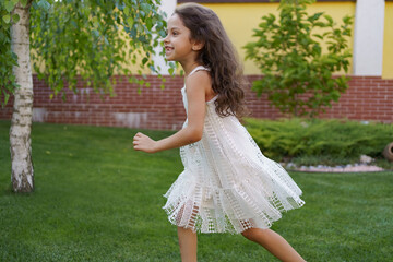 Fototapeta na wymiar Profile portrait of beautiful little girl with smiling face and long brunette wavy hair running happy barefoot in the backyard on the grass