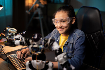 Portrait of girl sitting at desk in the evening, finishing soldering robot which is school project, child is watching tutorials, instructions on laptop, learning step by step how to fix electronics
