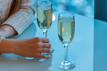 A pair of slender glass goblets with white sparkling wine stands on a glossy table, a woman's hand touches one of the glasses