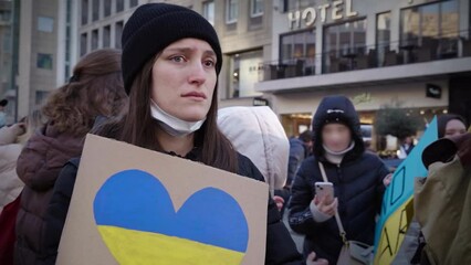 A pretty young Ukrainian woman holds a cardboard with the Ukrainian flag in the shape of a heart. She protests crying against the war in Ukraine. (2) - Powered by Adobe