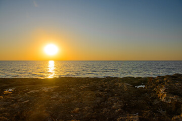 The setting sun on the horizon above the surface of a calm sea with a rocky shore