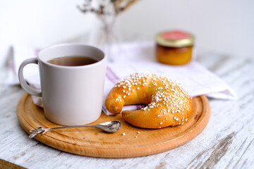 Bagel bun on a wooden round plate with a cup of tea and spoon on a table. Cozy breakfast. Sweet organic plant-based traditional bakery. Tea ceremony. Modern neutral light food photo template. 