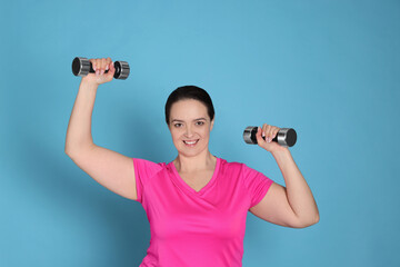 Fototapeta na wymiar Happy overweight woman doing exercise with dumbbells on light blue background