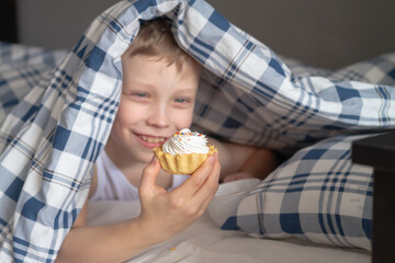 A Caucasian boy is eating a delicious cream cake lying under a warm blanket in a blue cage on the bed