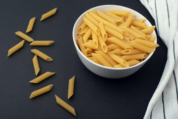 A bowl of raw pasta on a dark background. Scattered pasta and a dishcloth. 