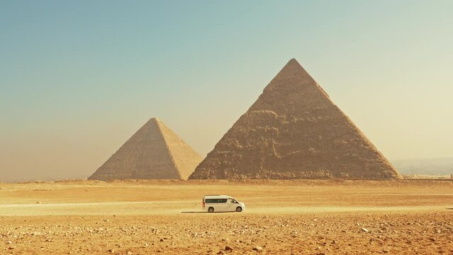 Bus carries tourists along sandy road look to pyramids on Giza Plateau near Cairo. Transport drives past necropolis, an Egyptian landmark. Minibus with travellers moving to ancient monuments