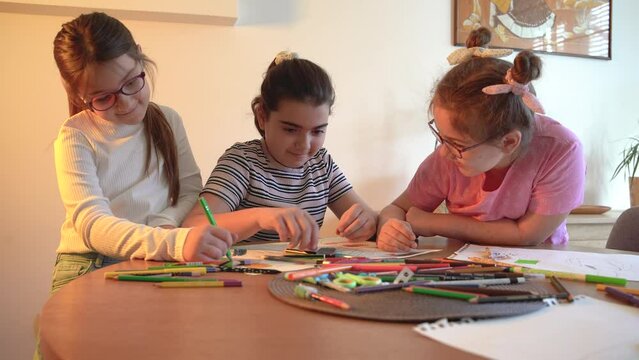 A group of school girls draw with color pencils. They're sitting at the table in the living room and having fun.